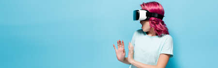 scared young woman with pink hair in vr headset gesturing on blue background, panoramic shot