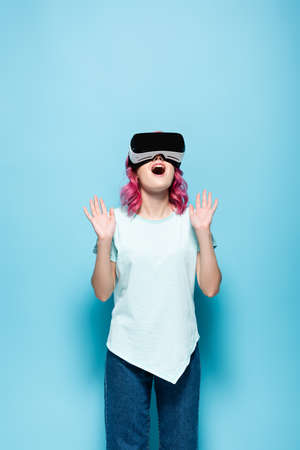 shocked young woman with pink hair in vr headset gesturing on blue background