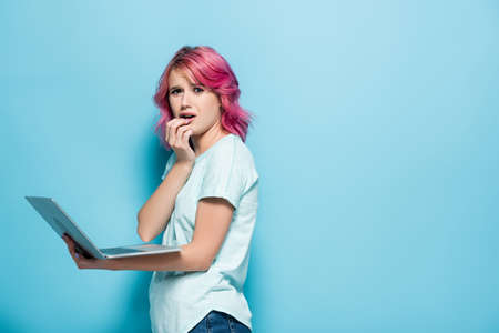 side view of worried young woman with pink hair holding laptop on blue backgroundの写真素材