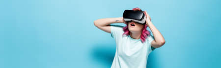 shocked young woman with pink hair in vr headset holding head on blue background, panoramic shot