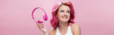 young woman with colorful hair holding headphones and smiling isolated on pink, panoramic shotの写真素材