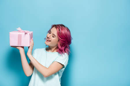 young woman with pink hair holding gift box with bow and smiling on blue background