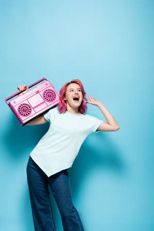 excited young woman with pink hair holding vintage tape recorder on blue backgroundの写真素材