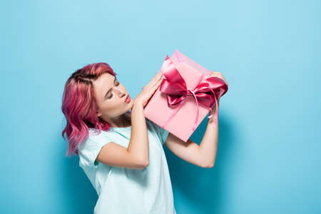 young woman with pink hair kissing gift box with bow on blue background