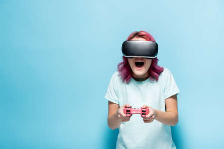 excited young woman with pink hair in vr headset holding joystick on blue background