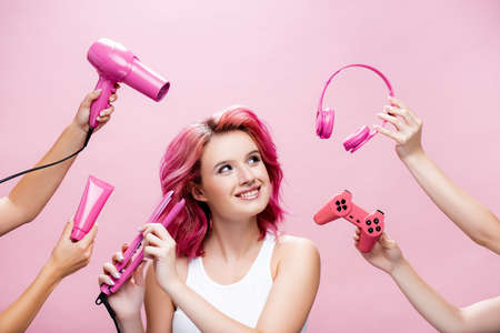 young woman with colorful hair using straightener near headphones, cosmetic cream, joystick and hairdryer in hands isolated on pinkの写真素材