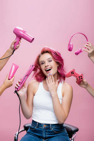 young woman with colorful hair near straightener, headphones, cosmetic cream, joystick and hairdryer in hands isolated on pinkの写真素材