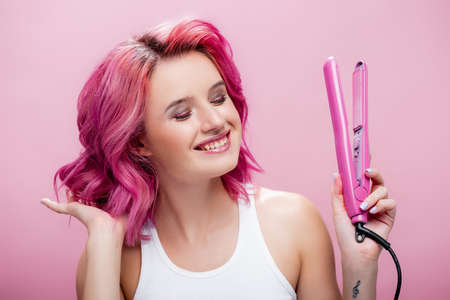 young woman touching colorful hair and holding straightener isolated on pinkの写真素材