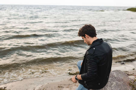 Man in leather jacket sitting on stone on sea coastの写真素材