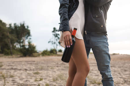 Cropped view of woman in dress and leather jacket holding bottle of wine near boyfriend on beach