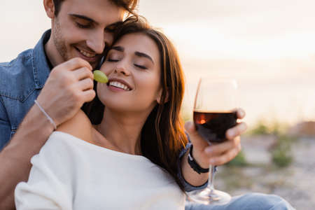 Selective focus of man feeding girlfriend with grape while holding glass of wine on beachの写真素材