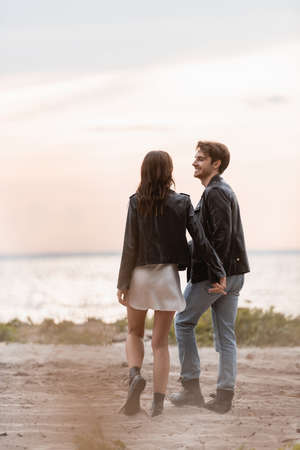 Selective focus of young couple in leather jackets holding hands while walking on beach at sunset