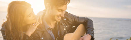 Panoramic shot of woman hugging boyfriend playing acoustic guitar during sunset on beach