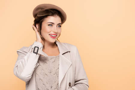 sensual woman in trench coat and beret looking away and touching hair isolated on peach