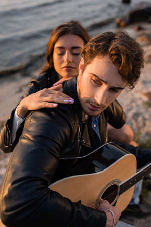 Selective focus of man playing acoustic guitar near woman on beach near sea at sunsetの写真素材