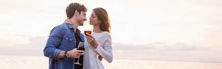 Panoramic orientation of woman hugging boyfriend while holding glass of wine near seaの写真素材