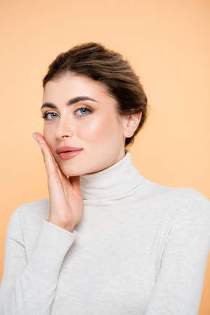 sensual woman in turtleneck touching face while looking at camera isolated on peach