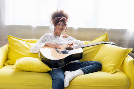 pleased woman holding acoustic guitar and sitting on sofa in living roomの写真素材