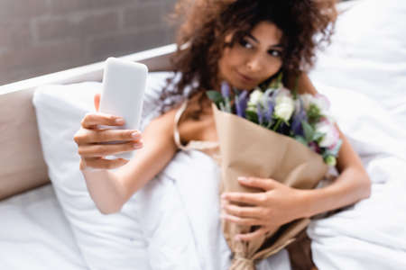 selective focus of curly woman taking selfie with flowers in bedroomの写真素材