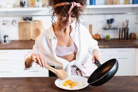 joyful woman holding frying pan and kitchen tongs while serving fried eggs on plateの写真素材