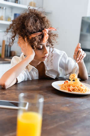 selective focus of displeased woman holding fork near plate with prepared spaghetti, glass of orange juice and smartphone on tableの写真素材