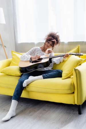 young curly woman playing acoustic guitar on sofa in living roomの写真素材