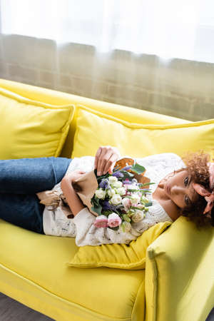 young curly woman lying on yellow sofa with flowers and looking awayの写真素材