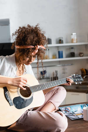 young curly woman playing acoustic guitar near cup on tableの写真素材