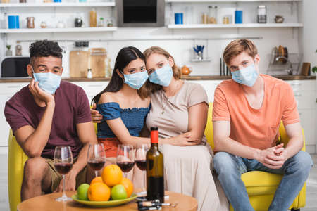 sad multiethnic friends in medical masks sitting at table with red wine and fruits in kitchenの写真素材