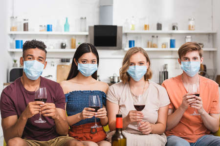 multicultural friends in medical masks holding glasses of red wine while looking at camera in...
