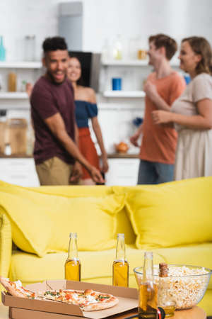 selective focus of beer and pizza on table near yellow sofa and multiethnic friends dancing on backgroundの写真素材