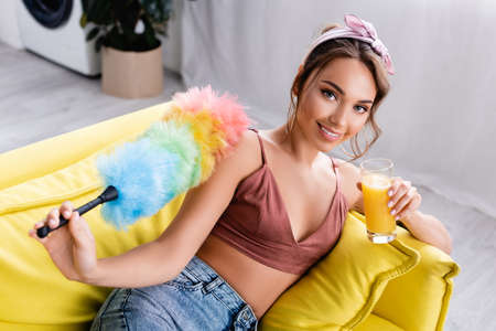 Selective focus of housewife holding dust brush and glass of orange juice on couchの写真素材
