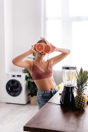 Selective focus of woman covering face with halves of grapefruit near blender on tableの写真素材