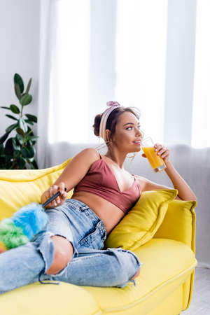 Selective focus of housewife drinking orange juice while holding dust brush on couch