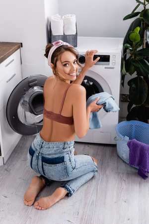 Back view of modern housewife looking at camera near clothes and washing machineの写真素材