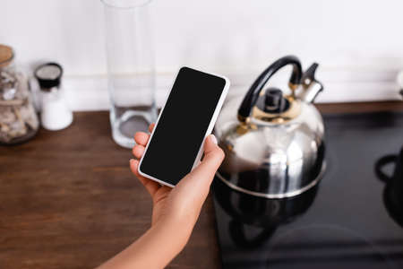 Cropped view of woman holding smartphone with blank screen near kettle on stoveの写真素材