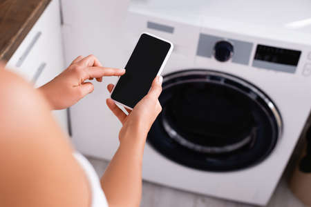 Cropped view of young woman using smartphone near automated washing machine