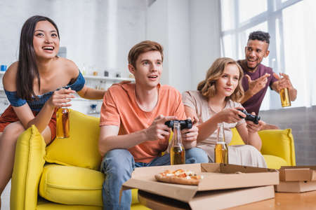 KYIV, UKRAINE - JULY 28, 2020: excited asian woman and african american man holding bottles of beer near friends playing video gameのeditorial素材