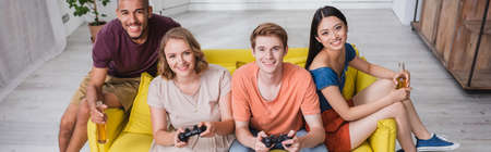 KYIV, UKRAINE - JULY 28, 2020: high angle view of multicultural friends holding beer and playing video game in kitchen, panoramic cropのeditorial素材
