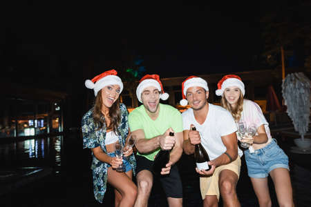 Excited friends with champagne bottles and glasses celebrating new year in swimming pool at nightの写真素材