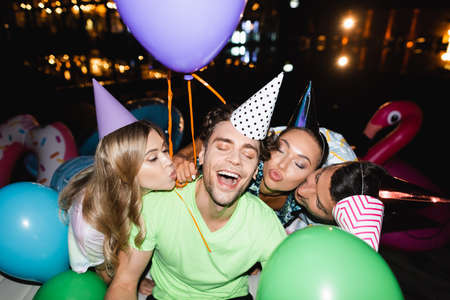 Selective focus of friends in party caps kissing man with balloons outdoors at nightの写真素材
