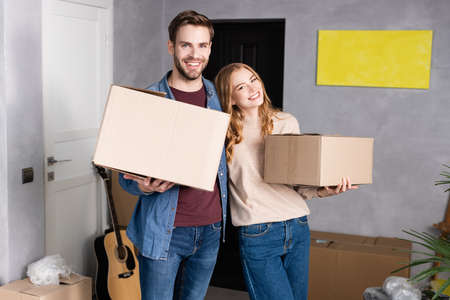 joyful man and woman holding carton boxes in new homeの写真素材