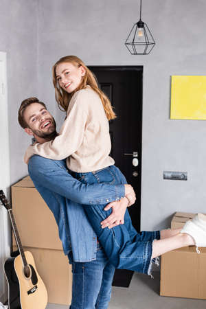 pleased man holding in arms woman near boxes and acoustic guitar, relocation concept
