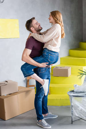 man holding in arms and looking at joyful girlfriend in new homeの写真素材