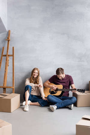 man playing acoustic guitar near pleased woman sitting near easel and boxes, moving conceptの写真素材