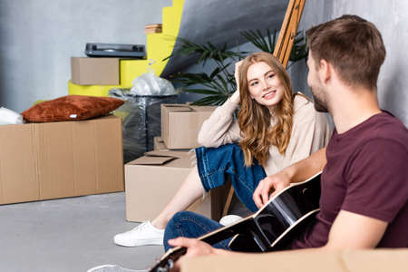pleased woman looking at man playing acoustic guitar near boxes, relocation conceptの写真素材