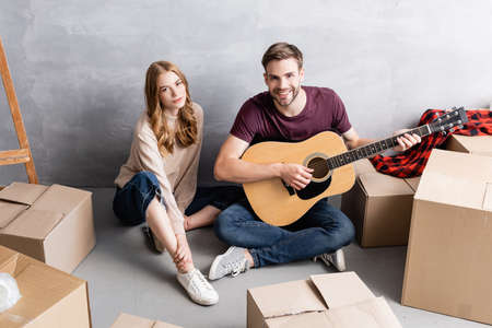 young woman sitting on floor with man playing acoustic guitar near boxes, relocation conceptの写真素材