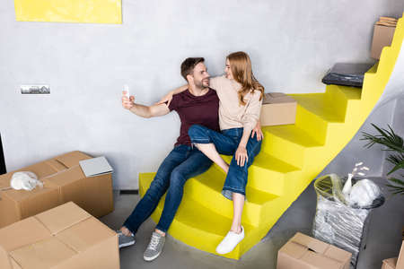 joyful couple sitting on yellow stairs and man taking selfie near carton boxes