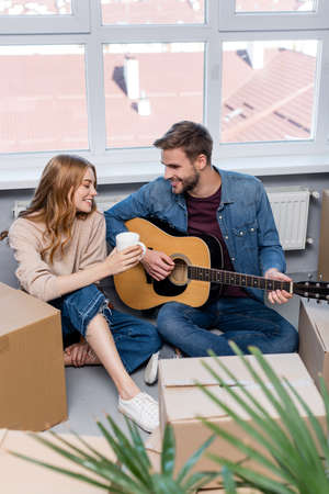 selective focus of young man playing acoustic guitar near woman with cup and carton boxesの写真素材
