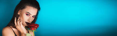 panoramic shot of young woman with dark makeup holding red rose on blue